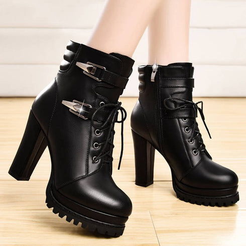 A pair of black high-heeled boots with lace-up closure and a thick heel, displayed against an indoor background.