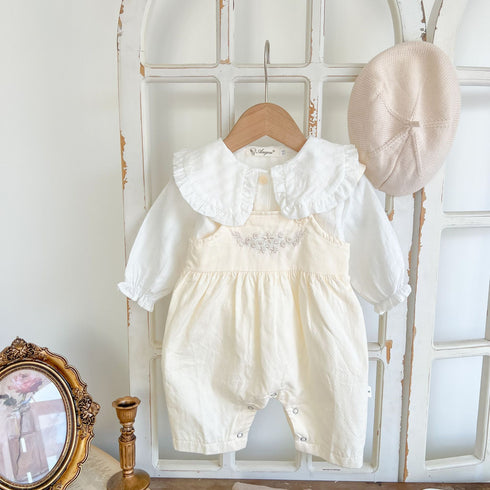 Baby girl's loose adjustable suspender romper in apricot and white, displayed on a wooden hanger with a beige hat and a mirror in the background.