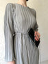 A woman wearing an elegant pleated bell sleeve mid-length grey dress with a mid-waist.