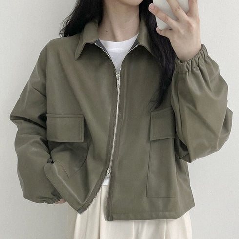 A woman wearing a long sleeve short coat in army green, with a relaxed fit, cropped jacket style, and a zipper down the center. She is also wearing a white top and beige skirt.