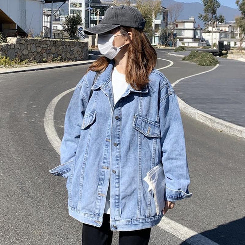 A lady wearing a loose denim jacket with a cap, mask, and sunglasses, standing on a street with a background of houses and a mountain.