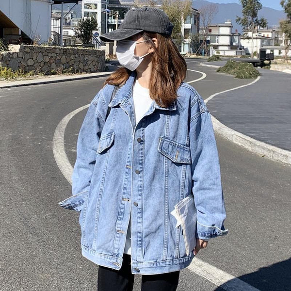 A lady wearing a loose denim jacket with a cap, mask, and sunglasses, standing on a street with a background of houses and a mountain.