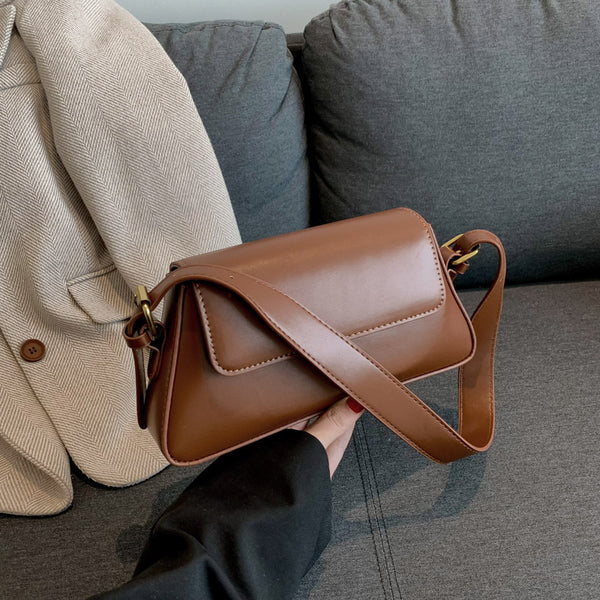 A hand holding a brown personalized messenger bag with adjustable strap, with a jacket and couch in the background.