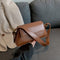 A hand holding a brown personalized messenger bag with adjustable strap, with a jacket and couch in the background.