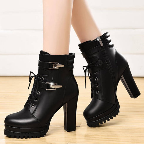 A pair of black high-heeled boots with lace-up closure and a thick heel, displayed against an indoor background.