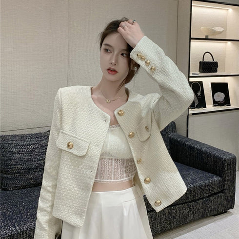 A woman standing in front of a white wall wearing a beige long sleeve tweed jacket with round neckline and single-breasted button details.