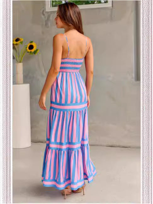 Back view of a woman standing in a sunny location wearing a pink and blue striped long dress with no sleeves and a square neckline.